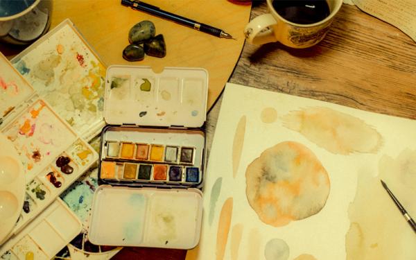 A watercolor palette and painting sit on a desk